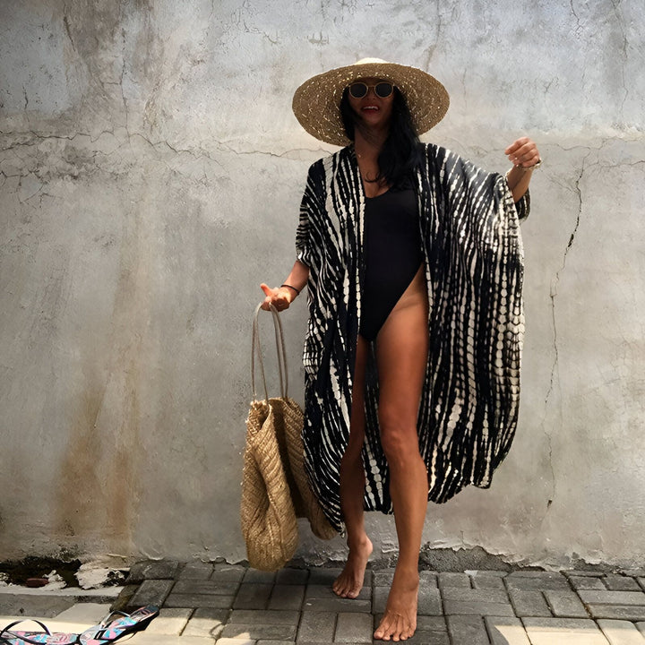 Femme portant un kimono noir et blanc à motifs, un maillot de bain noir, un chapeau de paille et des lunettes de soleil. Elle tient un sac de paille. Vente de kimonos.