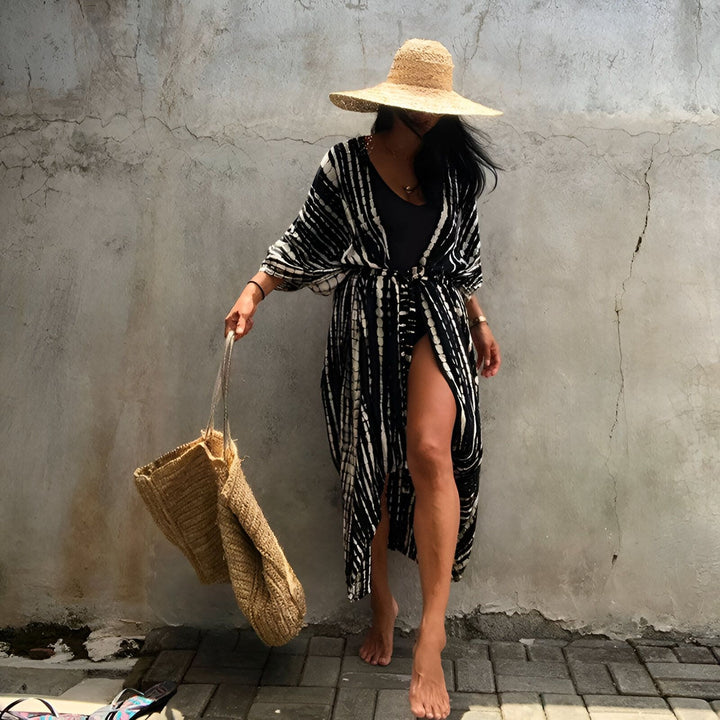 Femme portant un kimono noir et blanc à rayures, un chapeau de paille et un sac en osier. Style décontracté et élégant. Découvrez notre collection de kimonos uniques pour un look tendance et confortable.