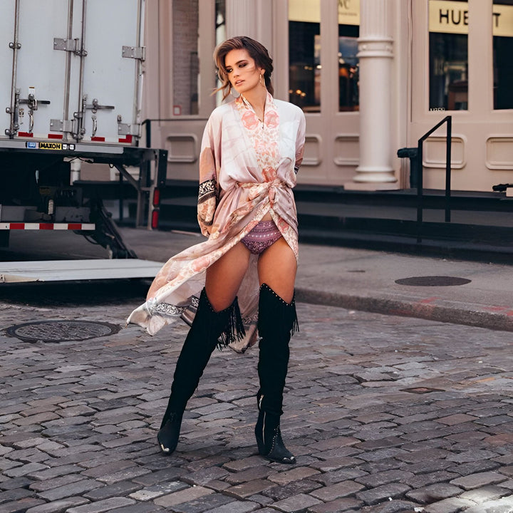 Femme portant un kimono rose à motifs floraux, noué à la taille, associé à des bottes hautes noires à franges. Mode kimono tendance. Découvrez notre collection de kimonos.