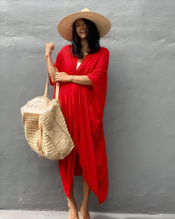 Femme souriante portant un kimono rouge vibrant, un chapeau de paille élégant et un sac tressé texturé. Look décontracté et tendance. Kimonos pour femmes disponibles.