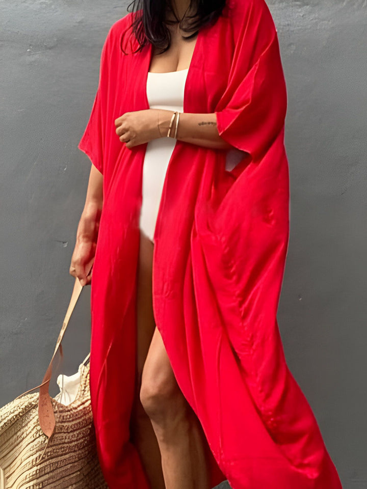 Femme portant un kimono rouge vif sur un maillot de bain blanc. Elle tient un sac de plage en paille. Vêtements de plage, kimono d'été. Découvrez notre collection de kimonos uniques.