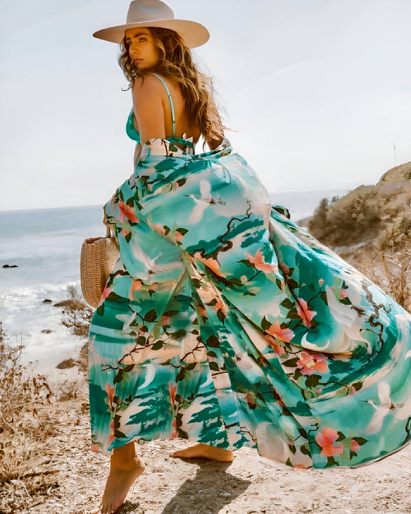 Femme portant un kimono fleuri turquoise et rose sur la plage. Chapeau beige et sac en paille. Mode kimono, vêtements de plage, style bohème. Découvrez notre collection de kimonos uniques.