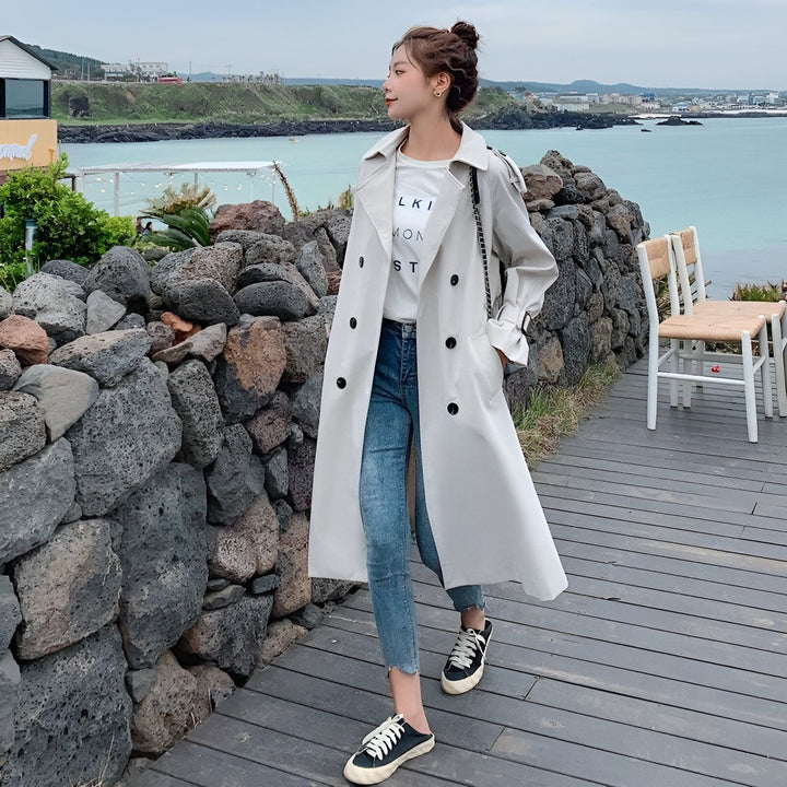 Femme élégante portant un kimono moderne long beige, jean et baskets, posant près de la mer. Kimonos de haute qualité à découvrir.