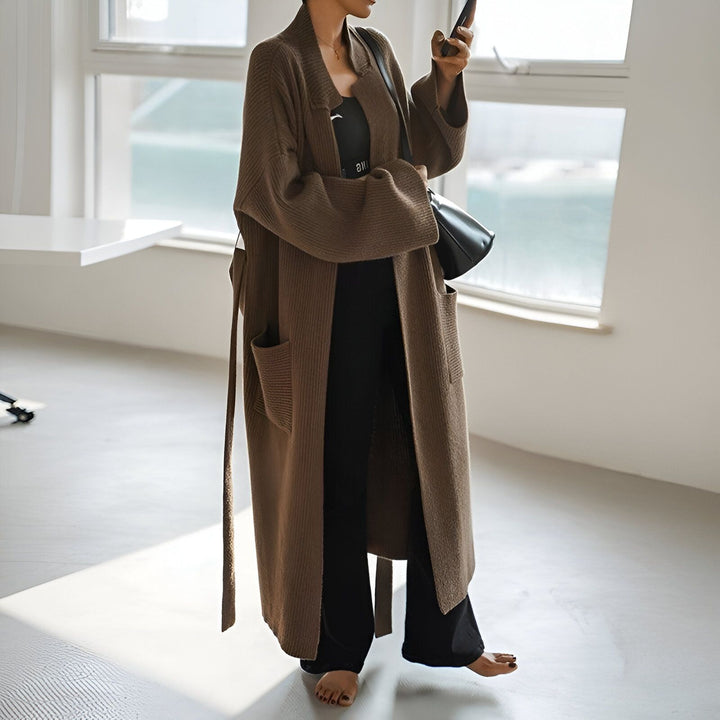 Femme portant un long kimono marron avec poches et ceinture. Elle tient un téléphone, avec un sac noir à l'épaule, près d'une fenêtre lumineuse. Vêtements de style kimono à la vente.