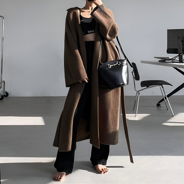 Femme portant un long kimono marron en tricot côtelé avec une ceinture, un soutien-gorge de sport noir et un pantalon noir. Sac à main noir sur l'épaule. Mode kimono.