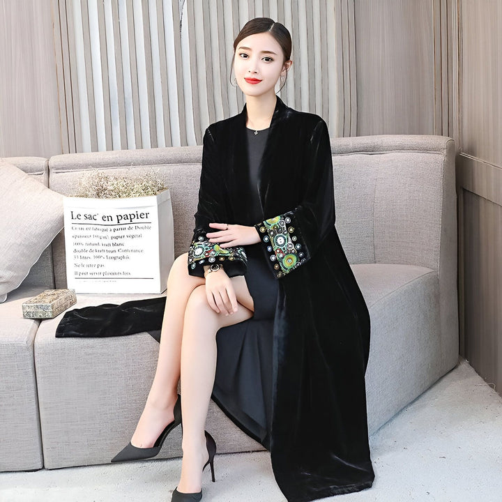 Femme élégante portant un kimono noir en velours avec broderies florales aux manches. Assise sur un canapé gris, talons hauts noirs. Vêtements kimono de haute qualité.