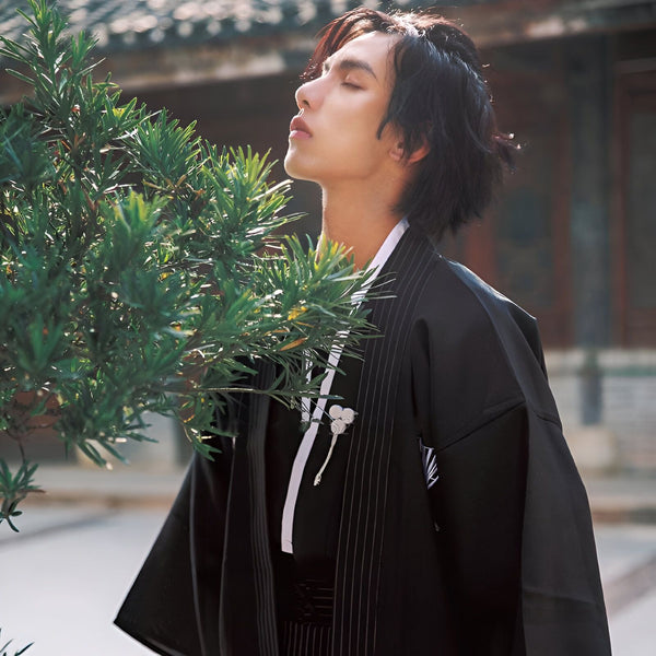 Homme portant un kimono japonais noir traditionnel avec des motifs blancs brodés, yeux fermés, appréciant la nature. Vêtements de style japonais en vente.