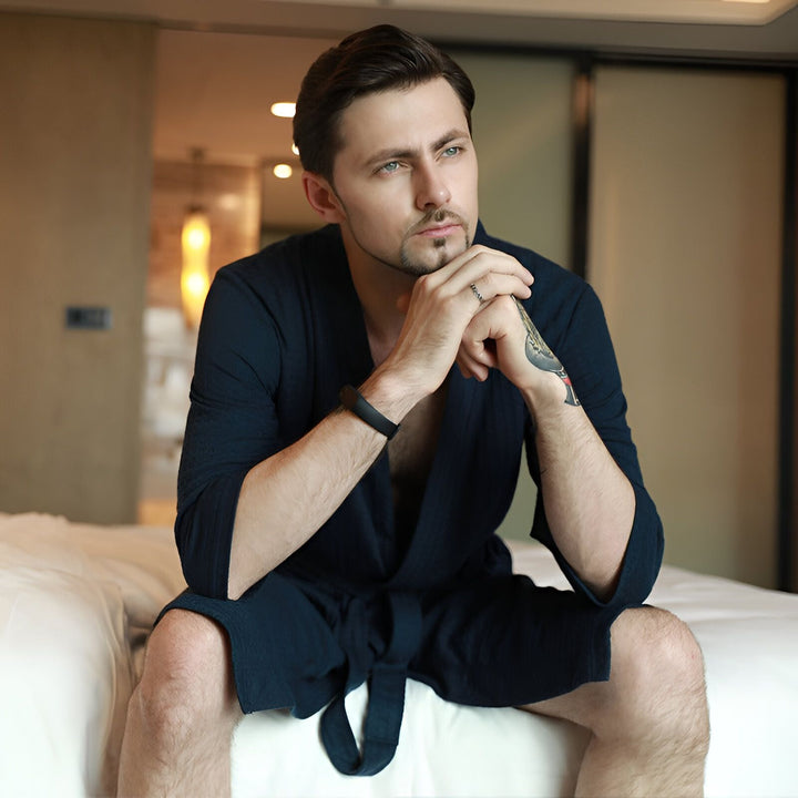 Homme portant un kimono bleu marine, assis sur un lit. Vêtements d'intérieur confortables et élégants. Kimonos pour hommes disponibles.