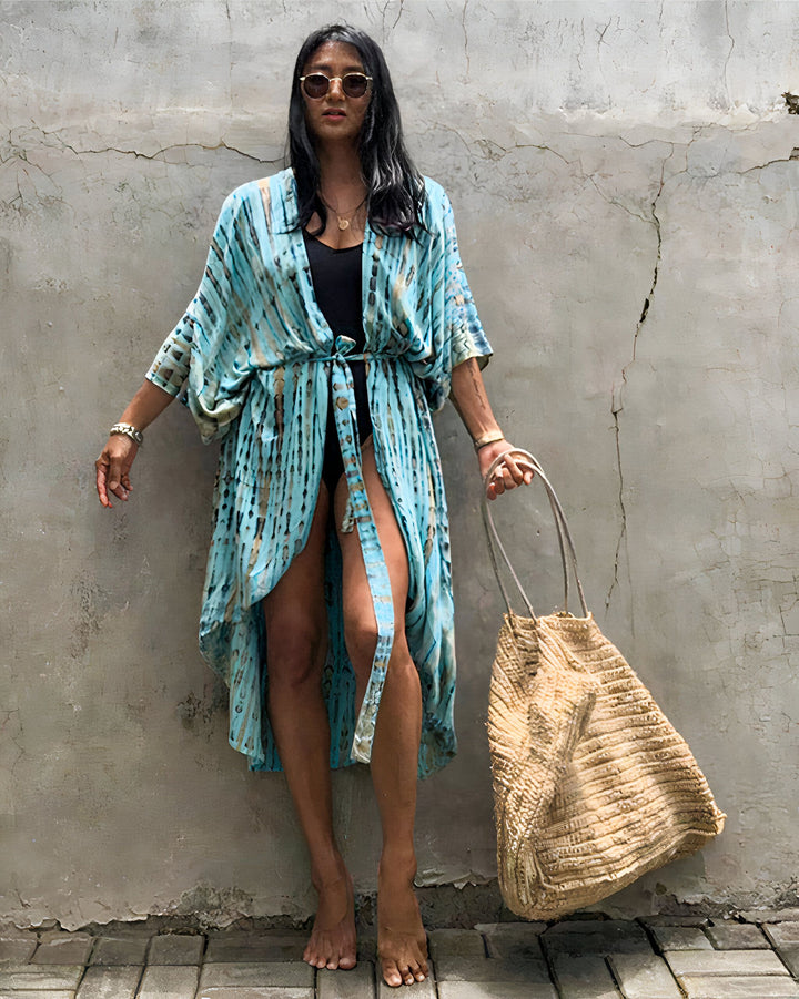 Femme portant un kimono bleu tie-dye avec un sac en paille. Kimono parfait pour la plage ou la maison. Découvrez notre collection de kimonos uniques et élégants.