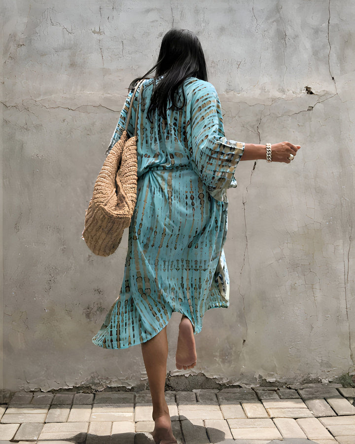 Femme vue de dos portant un kimono turquoise imprimé, un sac de paille et un bracelet. Kimonos tendance en vente dans notre boutique en ligne. Style décontracté et élégant.