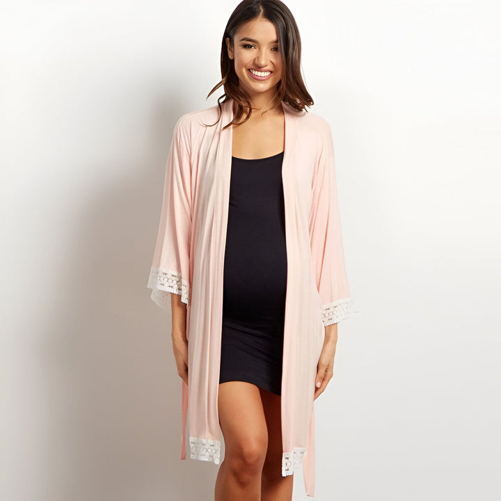 Femme enceinte souriante portant un kimono rose clair avec détails en dentelle blanche. Vêtements de maternité confortables. Kimonos pour femmes enceintes disponibles à l'achat.