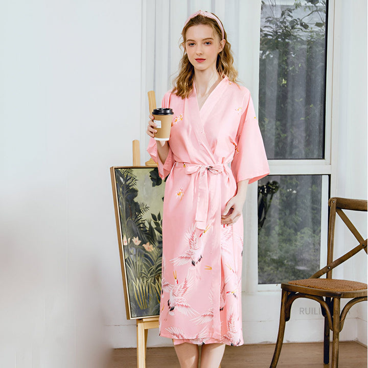Femme portant un kimono rose avec motifs de grues, tenant une tasse de café. Vente de kimonos en ligne. Style japonais traditionnel et moderne. Idée cadeau pour elle.