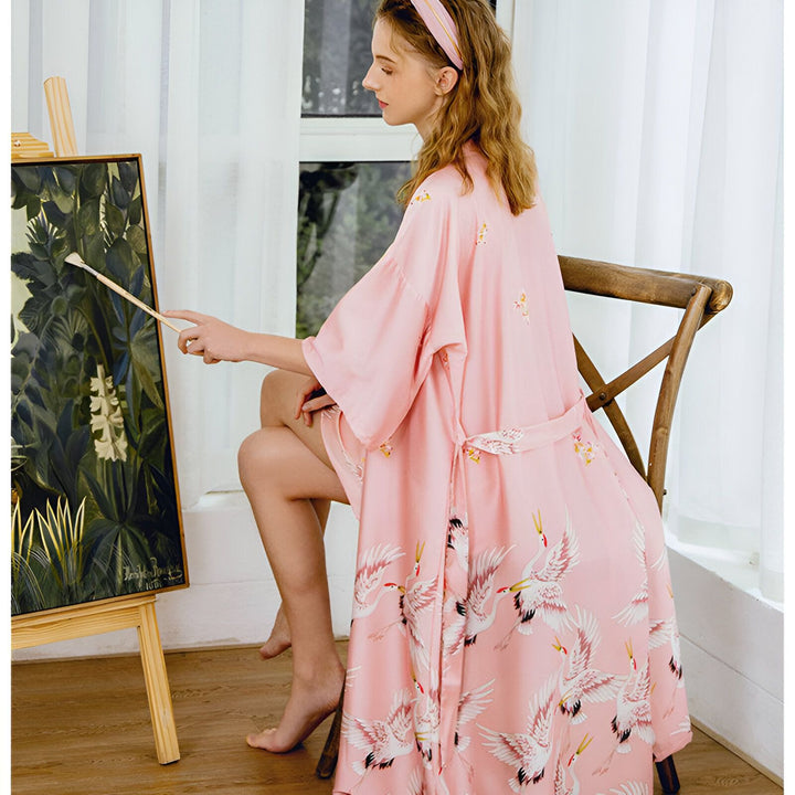 Femme peignant, portant un kimono rose à motifs de grues. Élégance et confort pour la maison. Découvrez notre collection de kimonos uniques.