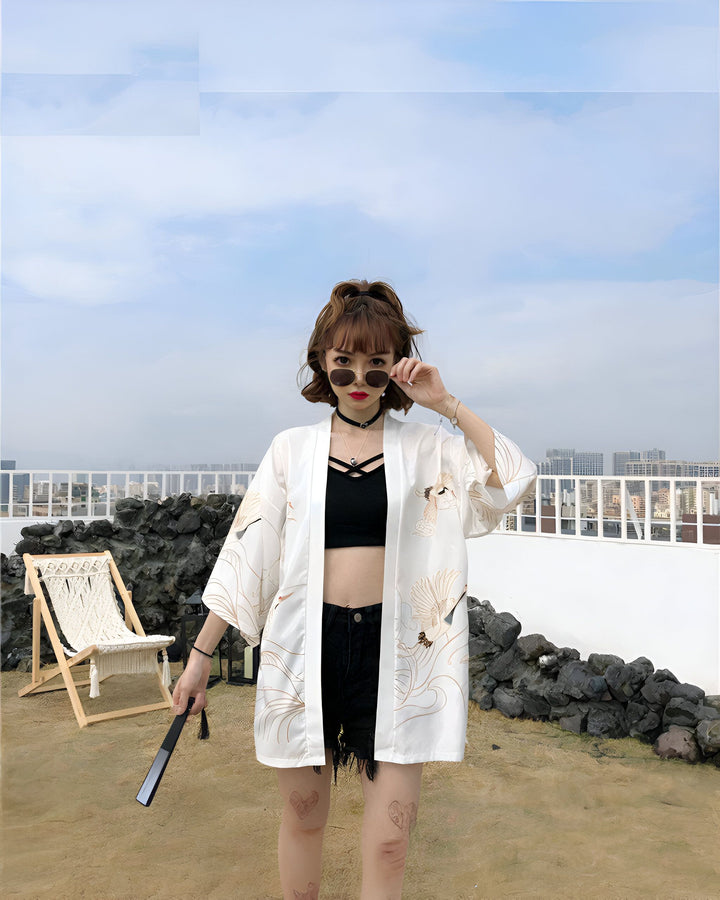 Jeune femme portant un kimono blanc à motifs de grues, avec un haut noir et un short en jean. Elle tient un éventail. Kimono tendance à acheter en ligne.