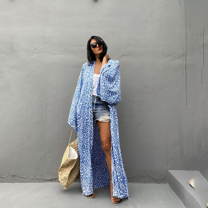 Femme portant un kimono long bleu à motifs, avec un short en jean et un débardeur blanc, et tenant un sac de plage. Vêtements de plage élégants. Kimonos disponibles à l'achat.