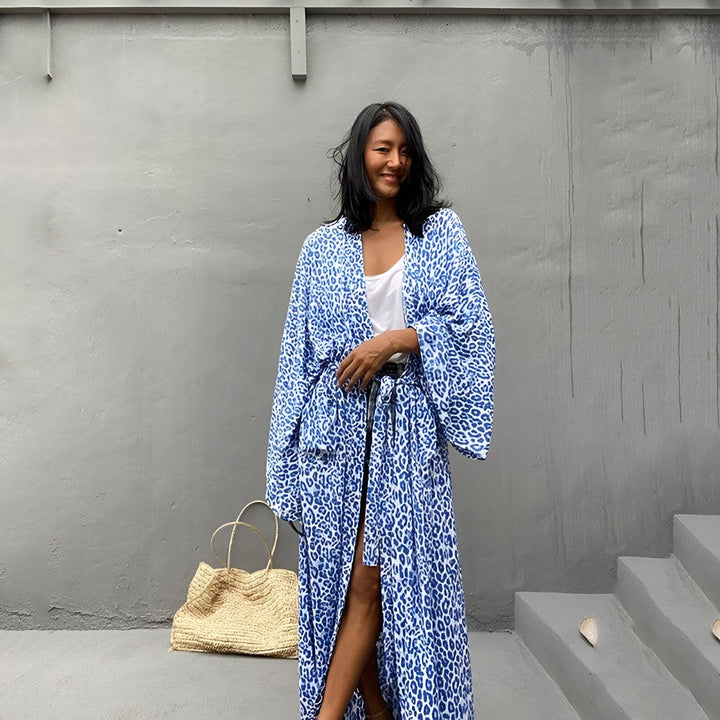 Femme souriante portant un kimono bleu à motif léopard. Look décontracté et élégant, parfait pour l'été. Sac en paille à côté. Découvrez notre collection de kimonos !