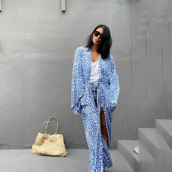 Femme portant un kimono bleu à motifs animaliers. Style décontracté avec un débardeur blanc et un short en jean. Sac en paille à côté. Kimonos tendance pour toutes les occasions.