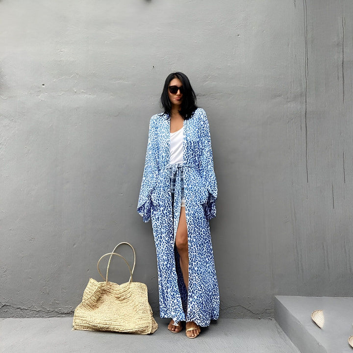 Femme portant un kimono long bleu à motifs léopard. Accessoires : lunettes de soleil, sac en paille. Collection de kimonos pour femmes.