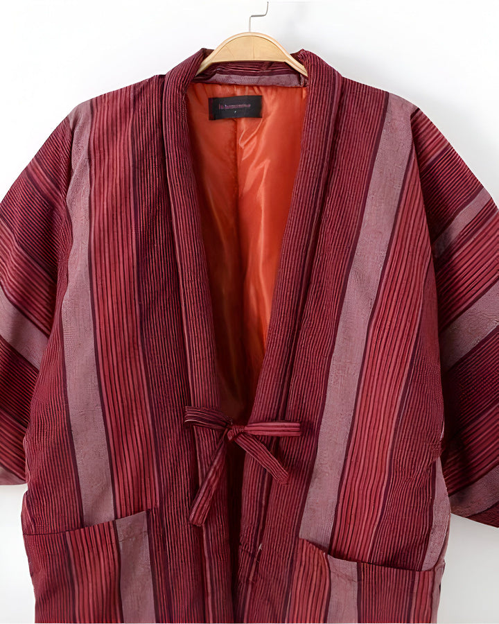 Kimono rayé rouge et bordeaux sur un cintre. Vêtement japonais traditionnel avec ceinture à nouer et poches. Parfait pour un look élégant et confortable.