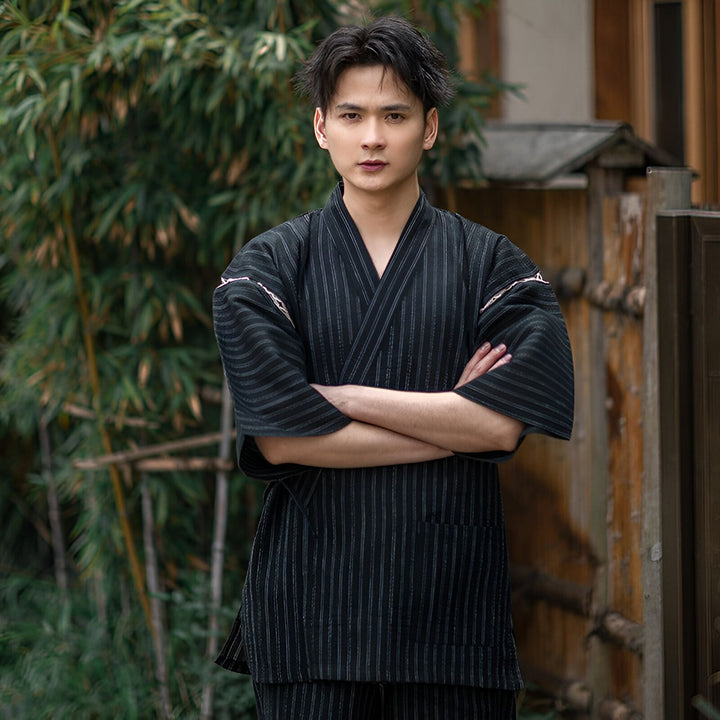 Homme portant un kimono japonais rayé noir et gris, les bras croisés devant un jardin. Vente de kimonos traditionnels et modernes.