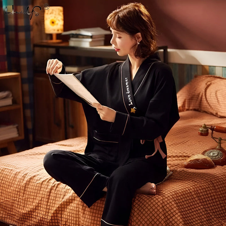 Femme assise sur un lit, lisant, portant un kimono noir élégant avec des détails roses. Vêtements de détente confortables, disponibles dans notre boutique de kimonos.
