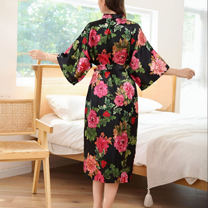 Femme portant un kimono noir à motifs floraux roses et verts devant une fenêtre. Kimono élégant et confortable pour la détente. Vêtements de nuit, peignoirs et robes de chambre japonaises.