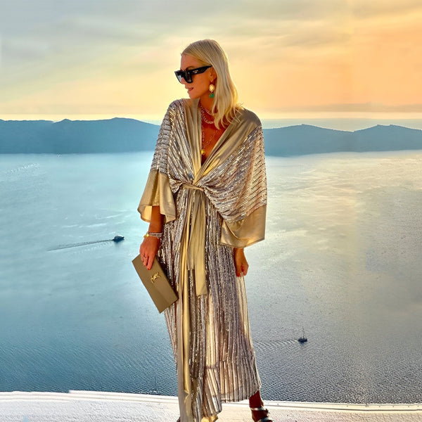 Femme élégante portant un kimono doré à motifs, avec vue panoramique sur la mer au coucher du soleil. Collection de kimonos chics et modernes.