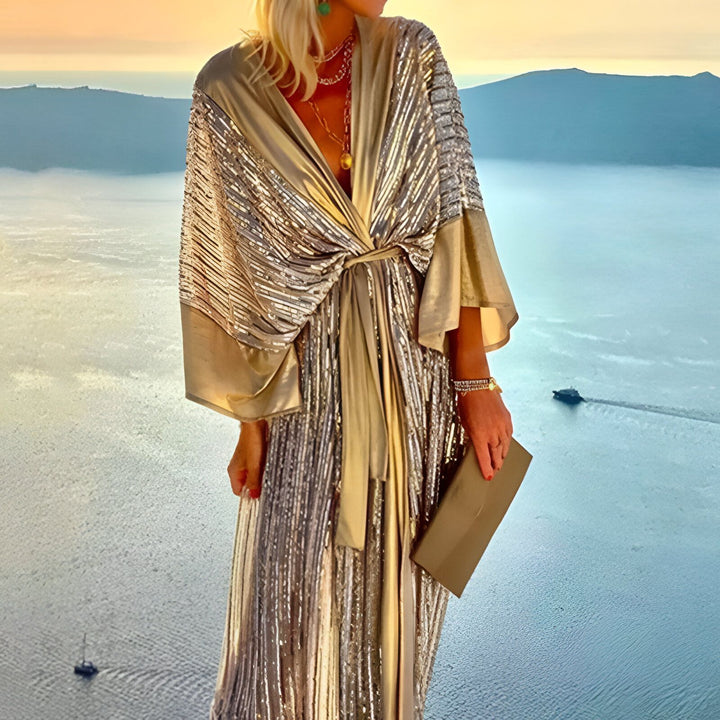 Femme portant un kimono long doré et argenté avec une ceinture, devant un paysage de mer et de montagnes au coucher du soleil. Kimonos de notre boutique.