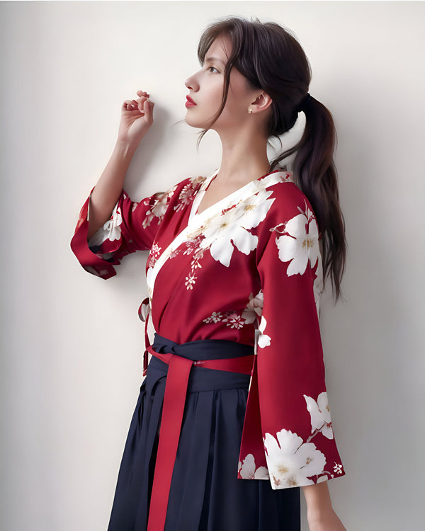 Jeune femme asiatique en tenue traditionnelle : kimono rouge fleuri et jupe bleu marine. Elle a les cheveux bruns attachés en queue de cheval et regarde sur le côté, appuiée contre un mur blanc. Mode asiatique, style vestimentaire.