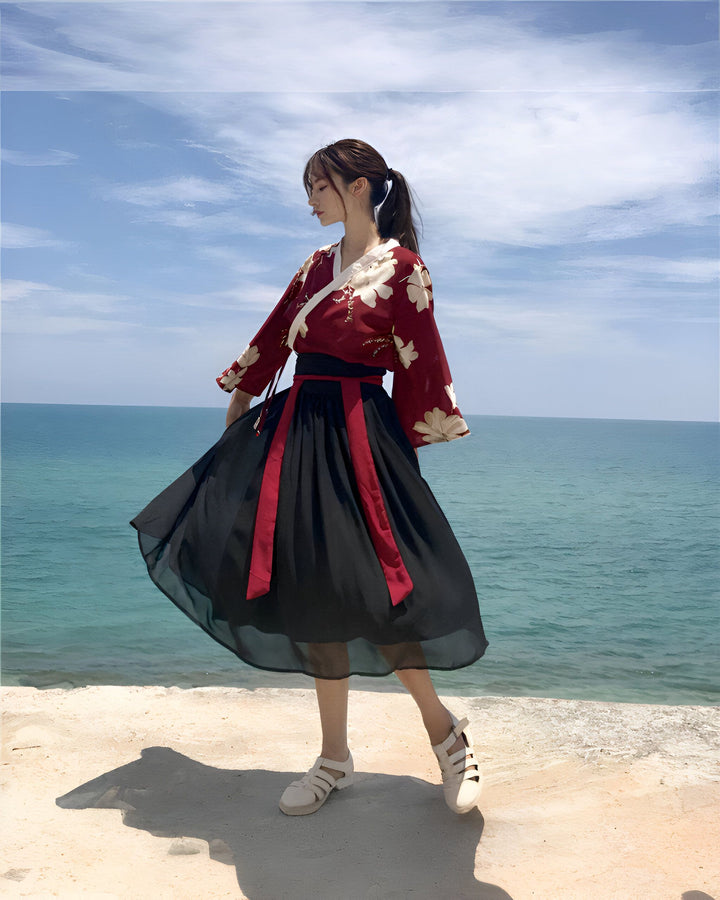 Jeune femme posant devant l'océan, vêtue d'une tenue traditionnelle chinoise rouge et noire avec des motifs floraux, une ceinture noire, et des sandales couleur crème. Cheveux attachés en queue de cheval, regardant vers le bas. Ciel bleu en arrière-plan.