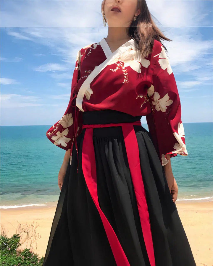 Femme portant un kimono rouge à fleurs blanches et une jupe noire avec des rubans rouges, devant un paysage de plage et d'océan. Style traditionnel asiatique revisité. Mode, voyage, culture.