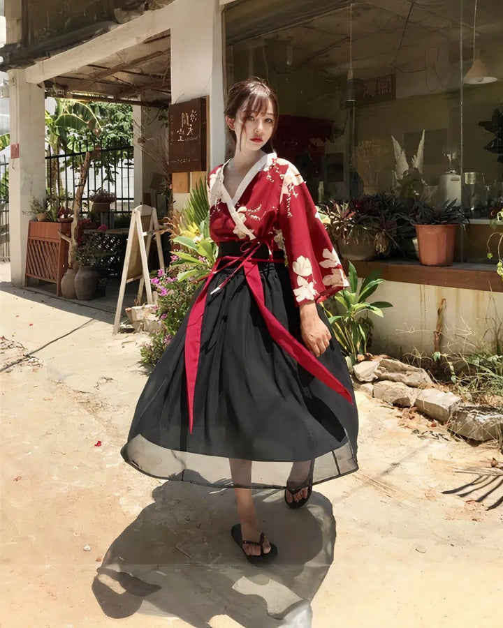 Jeune femme portant un kimono rouge à motifs floraux blancs et une jupe noire ample, ceinturée de rubans rouges. Elle pose devant une boutique avec des plantes. Mode asiatique, style traditionnel.