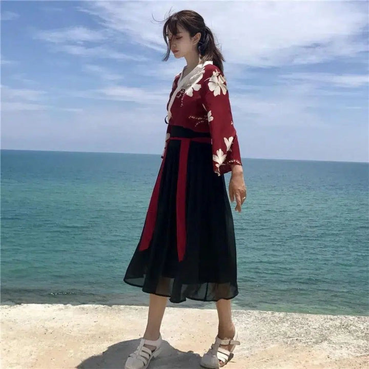 Jeune femme élégante en tenue d'inspiration asiatique, veste rouge à fleurs blanches et jupe noire, posant devant l'océan bleu. Mode estivale, style traditionnel revisité, look féminin et décontracté.
