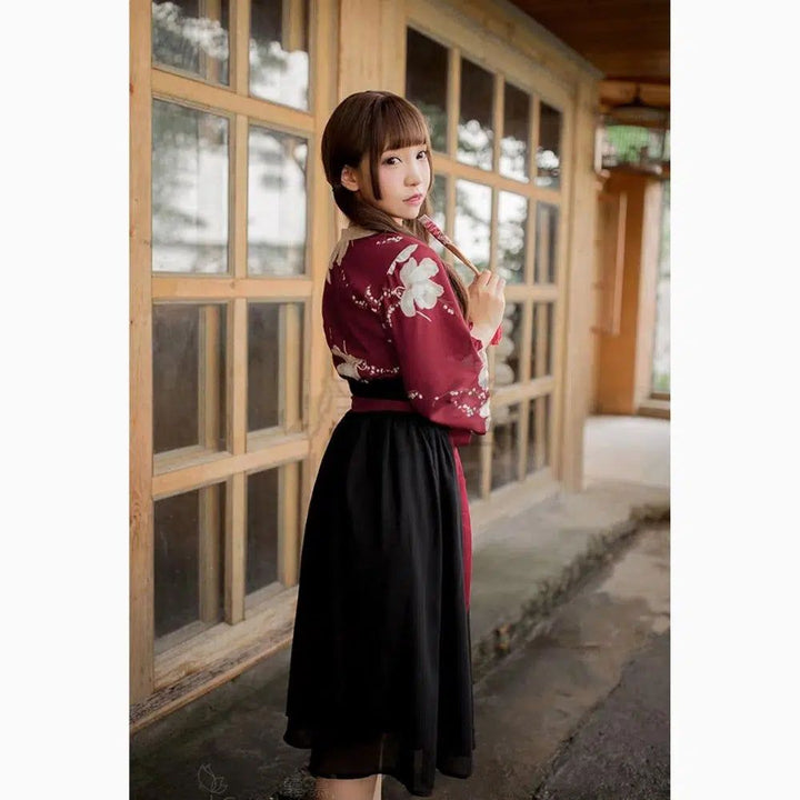Jeune femme en kimono bordeaux avec motifs floraux blancs, jupe noire. Portrait devant une maison traditionnelle japonaise en bois, expression douce. Mode asiatique.