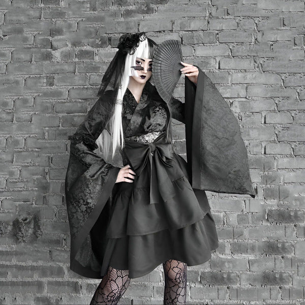 Femme gothique en kimono noir avec un éventail devant un mur de briques. Kimono à manches larges, robe à volants, voile sur la tête. Vêtements japonais alternatifs et uniques. Boutique de kimonos gothiques en ligne.