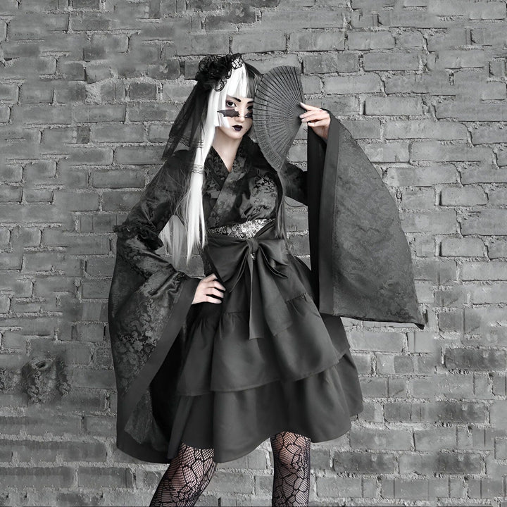 Femme gothique en kimono noir avec un éventail devant un mur de briques. Kimono à manches larges, robe à volants, voile sur la tête. Vêtements japonais alternatifs et uniques. Boutique de kimonos gothiques en ligne.