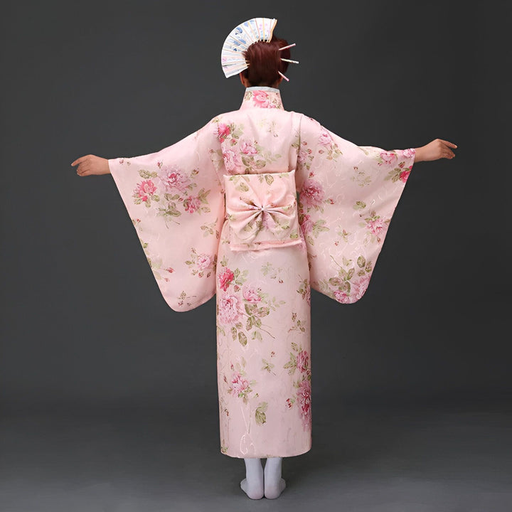 Femme portant un kimono rose à motifs floraux, vue de dos. Kimono traditionnel japonais, obi décoratif et éventail. Découvrez notre collection de kimonos uniques.