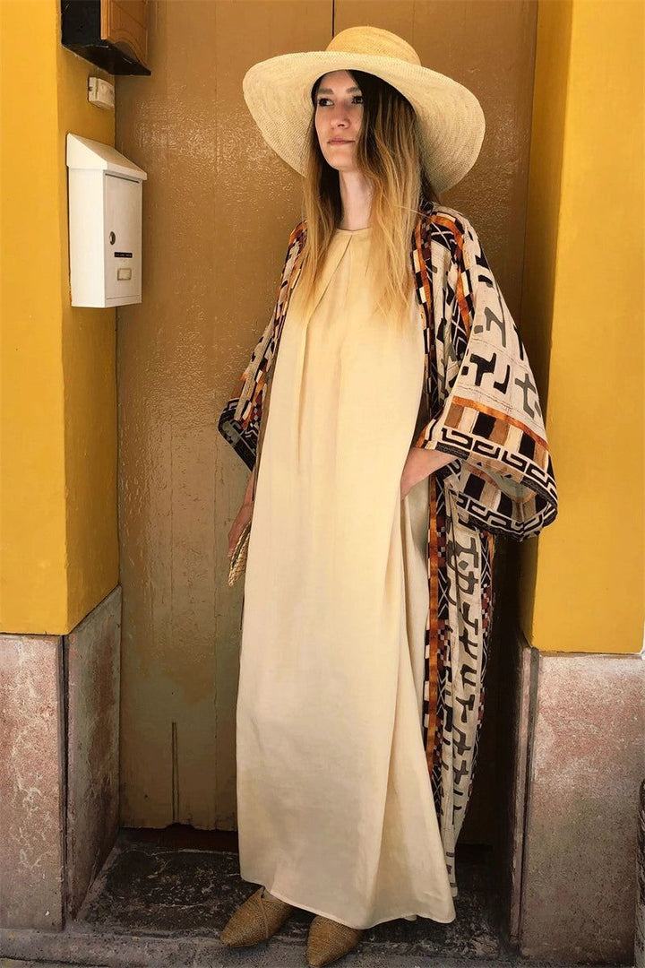 Femme portant un kimono à motifs et un chapeau de paille, adossée à un mur jaune. Découvrez notre collection de kimonos uniques.