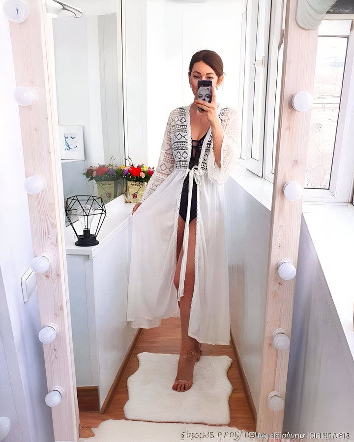 Femme en kimono blanc dentelle devant un miroir éclairé. Elle porte un maillot de bain noir et prend un selfie. Kimonos élégants et confortables disponibles.