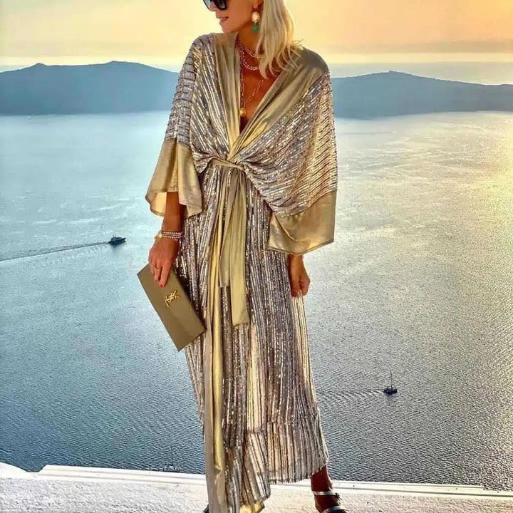 Femme portant un kimono long doré et argenté à paillettes. Vente de kimonos chics pour femmes. Kimono de soirée élégant avec ceinture. Accessoires de mode : bijoux, pochette. Arrière-plan : vue sur mer au coucher du soleil.