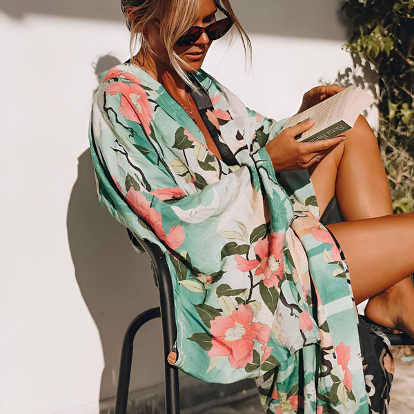 Femme lisant, portant un kimono fleuri turquoise et rose. Kimonos bohèmes pour femmes. Détente au soleil en kimono. Mode estivale en kimono fleuri.