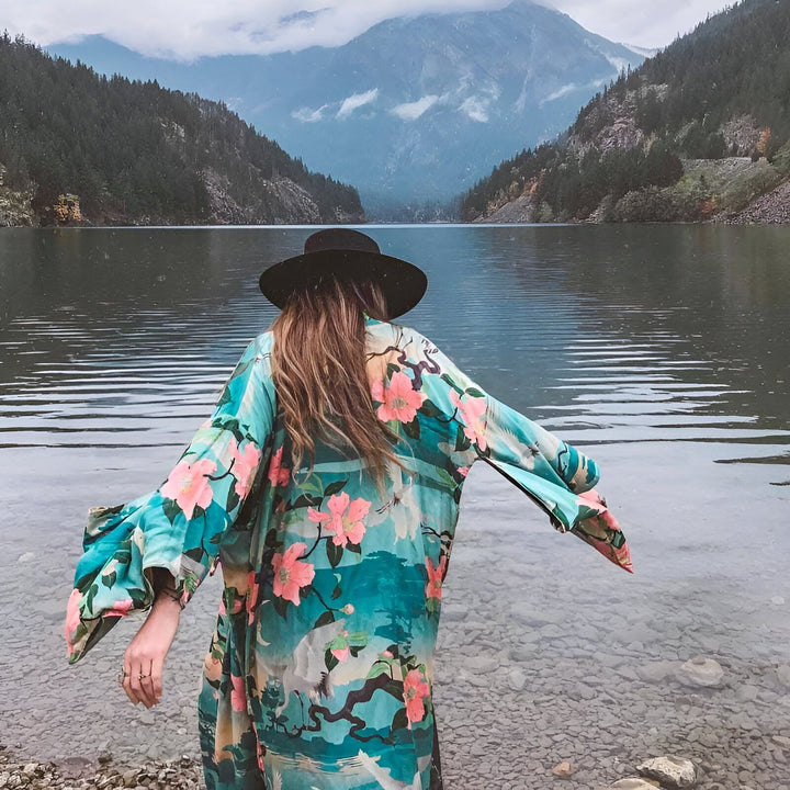 Femme vue de dos portant un kimono fleuri devant un lac et des montagnes. Kimono turquoise et rose, chapeau noir. Vêtements de style kimono.
