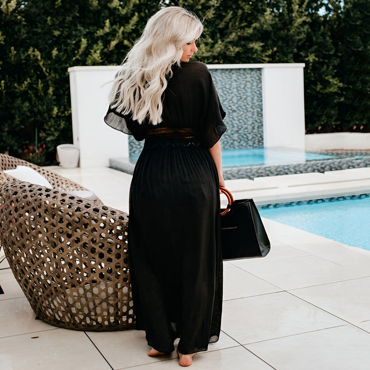 Femme blonde portant un kimono noir long près d'une piscine. Vue de dos, elle tient un sac à main noir. Kimono élégant et décontracté pour la plage ou la piscine. Découvrez notre collection de kimonos.