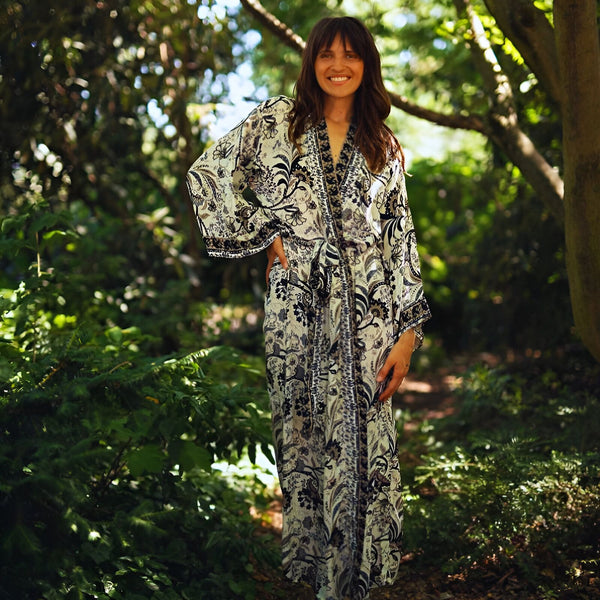 Femme souriante portant un kimono long à motifs floraux dans un cadre naturel luxuriant. Kimonos élégants disponibles à l'achat.