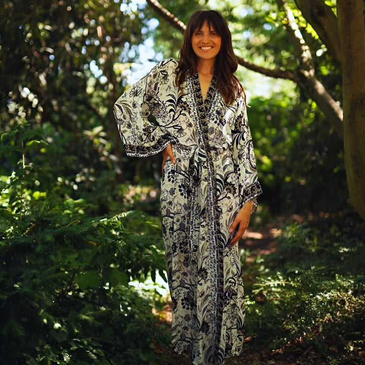 Femme souriante portant un kimono long à motifs floraux dans un cadre naturel luxuriant. Kimonos élégants disponibles à l'achat.