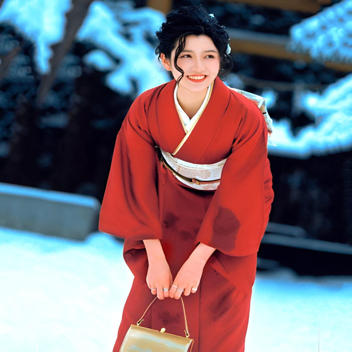 Jeune femme souriante en kimono rouge traditionnel, tenant un sac à main. Élégance japonaise et beauté en hiver. Découvrez notre collection de kimonos authentiques.