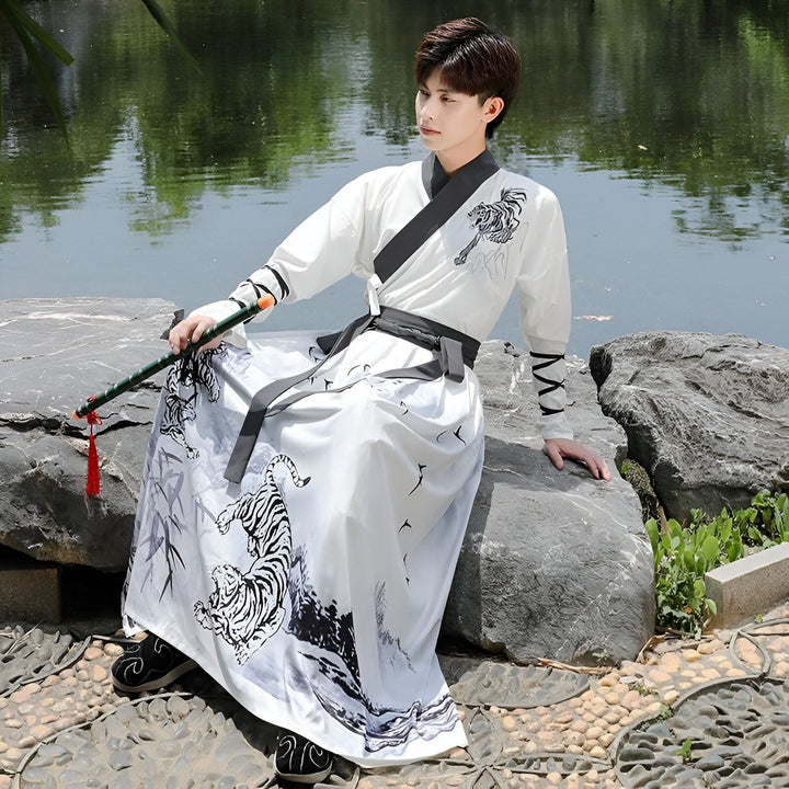 Un jeune homme en tenue traditionnelle chinoise blanche avec des motifs de tigres, assis sur une pierre près de l'eau. Il tient un bâton de bambou. Le paysage est paisible et serein.