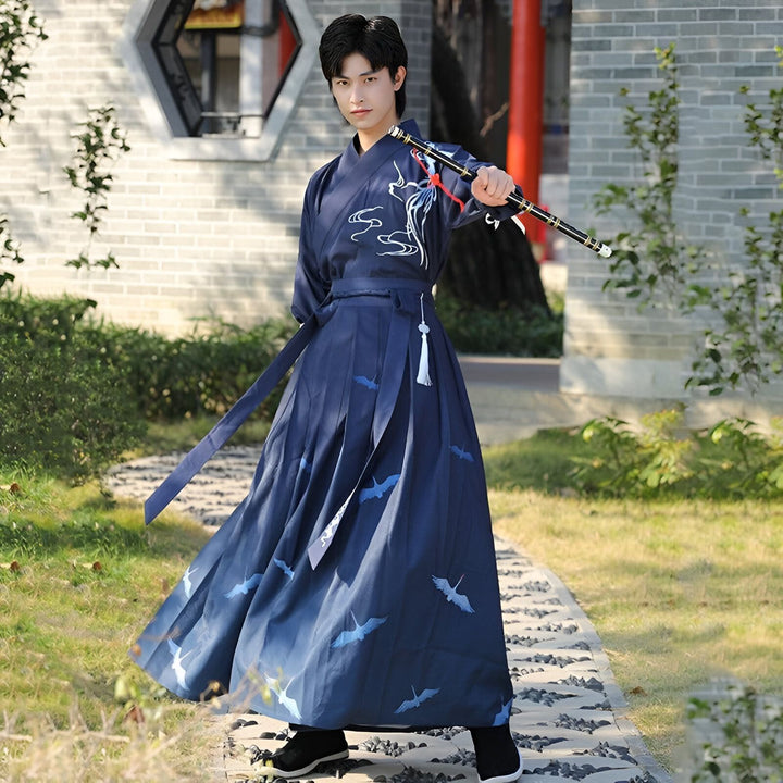 Un jeune homme en habit traditionnel bleu, tenant une flûte, pose dans un jardin. La tenue est ornée de motifs de grues et de nuages stylisés.
