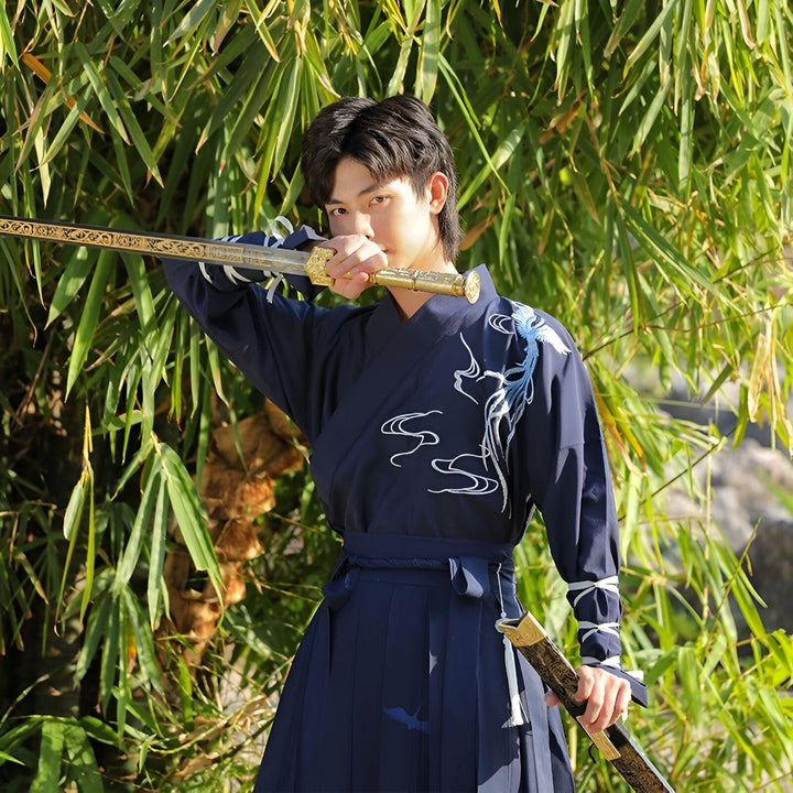 Jeune homme en costume traditionnel asiatique bleu marine, tenant un sabre doré devant un fond de bambous. Vêtement brodé de motifs de dragon. Accessoire de samouraï ou guerrier historique. Culture asiatique et art martial.