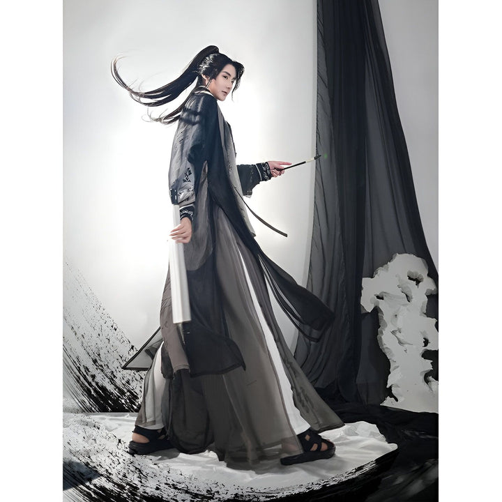 Homme en costume traditionnel asiatique. Il porte une longue robe fluide noire et grise, les cheveux attachés, et tient une baguette. Le fond est décoré de voilages noirs et blancs.